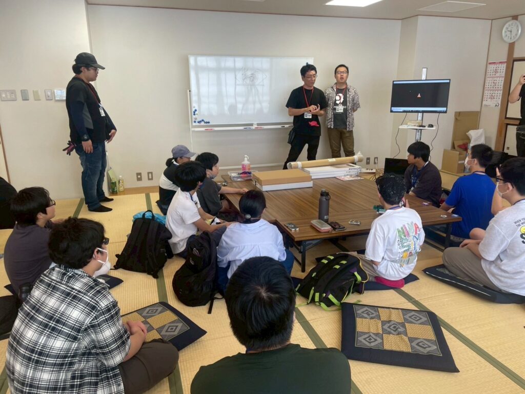 畳の部屋で、座布団に座った大勢の若者たちが、ホワイトボードの前に立つ講師の話を真剣な表情で聞いている様子を室内の後方から写した写真