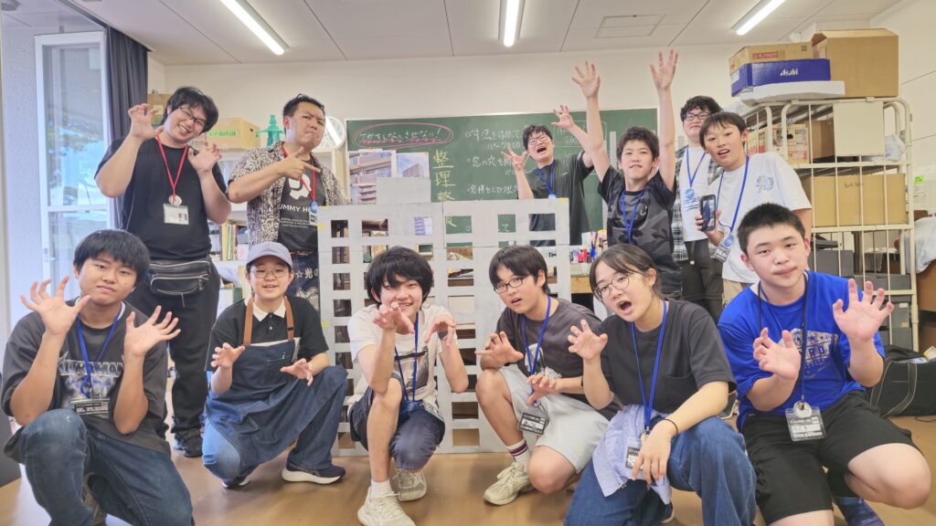 制作工程が書かれた黒板のある実習室で、格子状の窓が並ぶ大きな建物の模型を囲み、多くの学生たちが怪獣のようなポーズをとりながら、賑やかで楽しそうな表情で集合写真に収まっている様子を捉えた写真