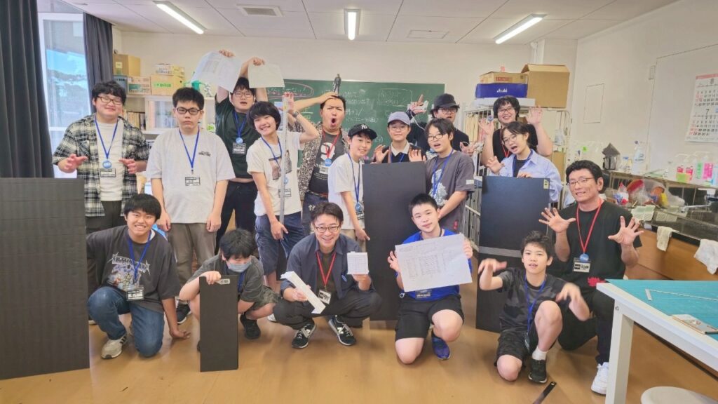 教室内で塾生たちが制作中の黒いボードや図面を手に掲げ達成感あるポーズで集合している記念写真