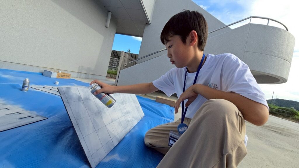 屋外で男性が青い養生シート上に置いた建物模型用パネルにスプレー塗装を施している写真