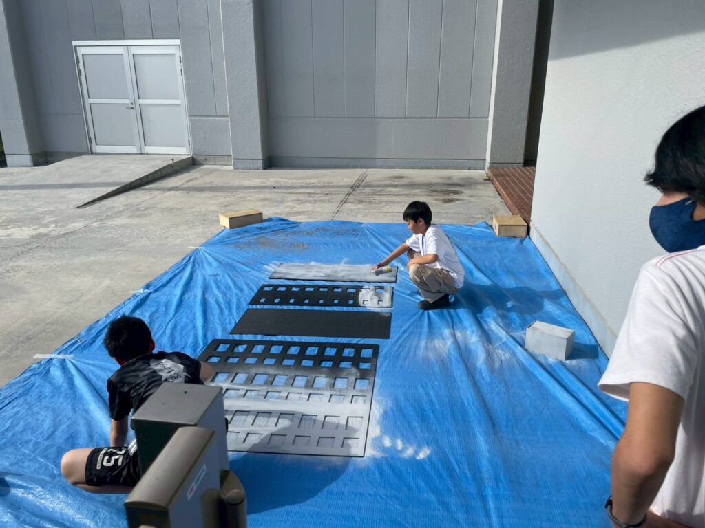 青い養生シート上で複数の塾生が建物模型用パネルにスプレー塗装を施している様子の写真