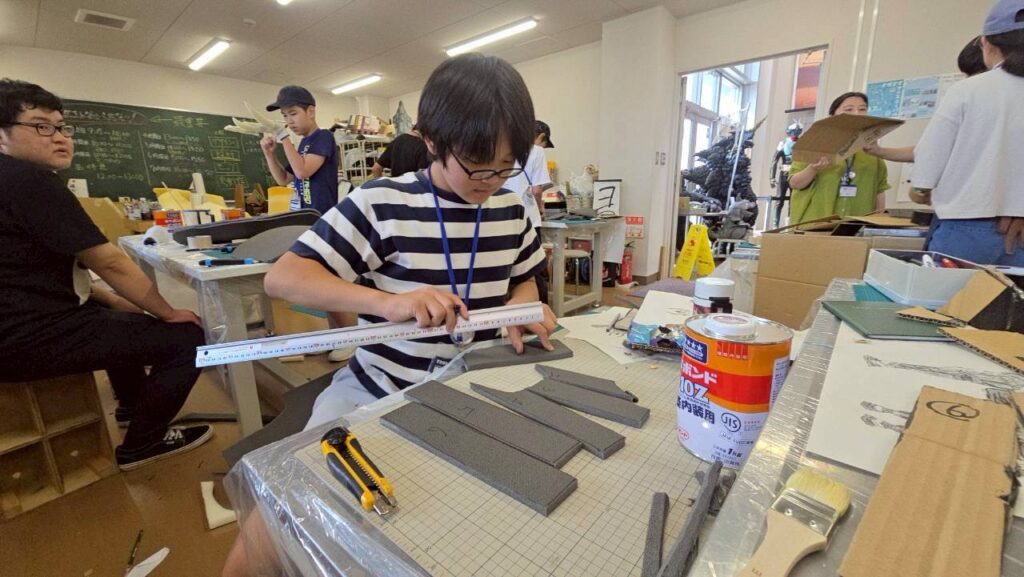 作業場の机で縞柄の服の塾生が長い定規で発泡素材の部材を測り周囲でも工作が進む様子を捉えた写真