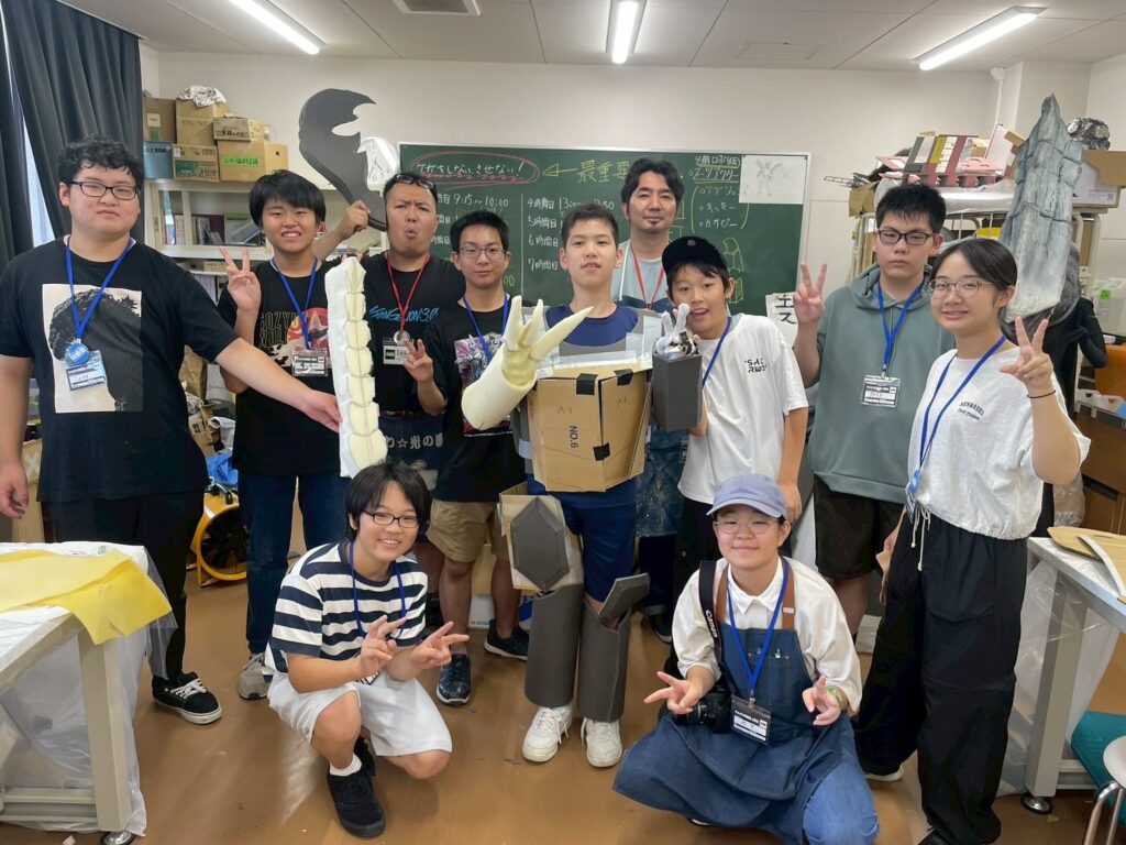 作業場で段ボール製の大型ロボットを中心に塾生たちが完成品を手に集まり笑顔でポーズを取って記念撮影する写真