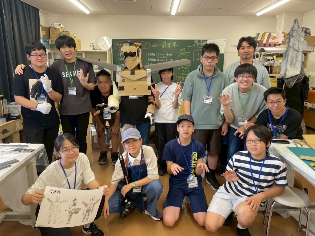 作業場で段ボール製ロボットを中心に塾生たちが完成品を手に集まりポーズを取って記念撮影する写真