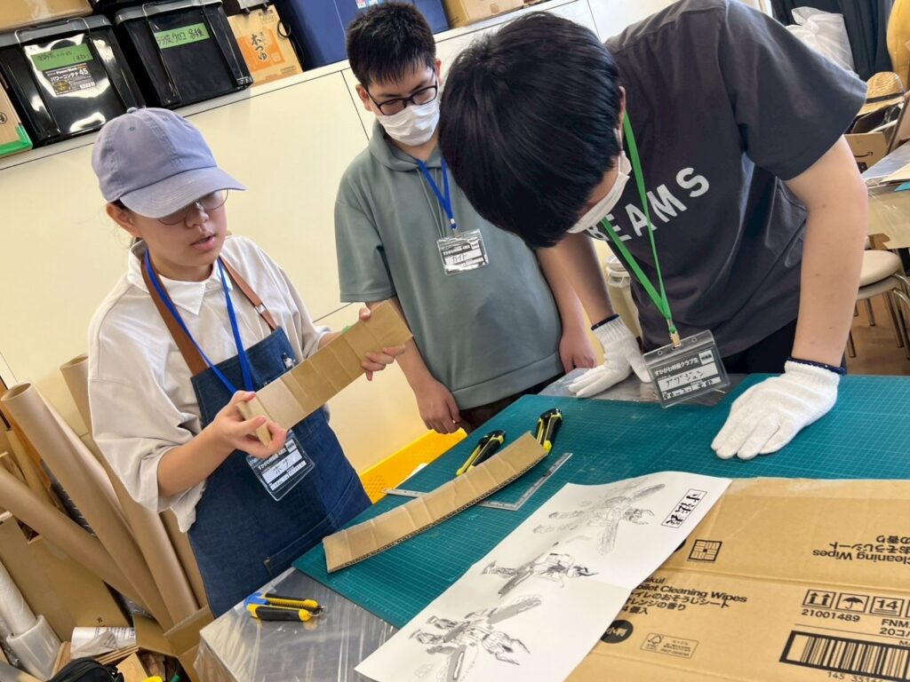 作業場の机で塾生たちが段ボール部材と図面を前に切断作業の手順を確認しながら工作を進めている写真