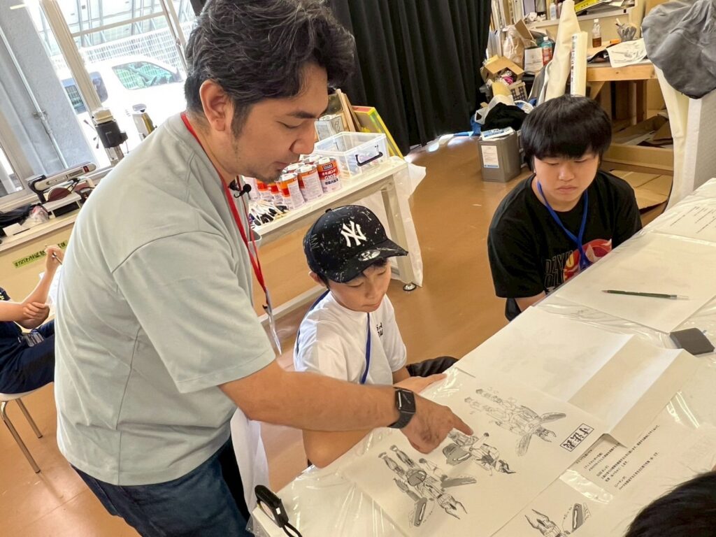 作業場の机で大人が図面を指し示し2人の子ども椅子に座り、静かな雰囲気の中で熱心に説明を受けている様子を捉えた写真