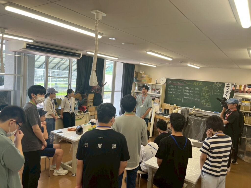 作業場の室内で塾生たちが黒板前に立つ講師を囲んで説明を受けている様子を広く捉えた写真