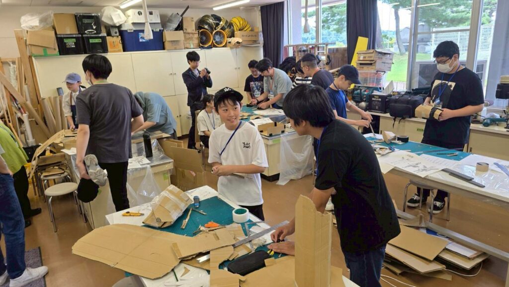 作業場の広い室内で塾生たちが段ボール材料と図面を使い複数の作業台で工作に集中して取り組む様子を捉えた写真