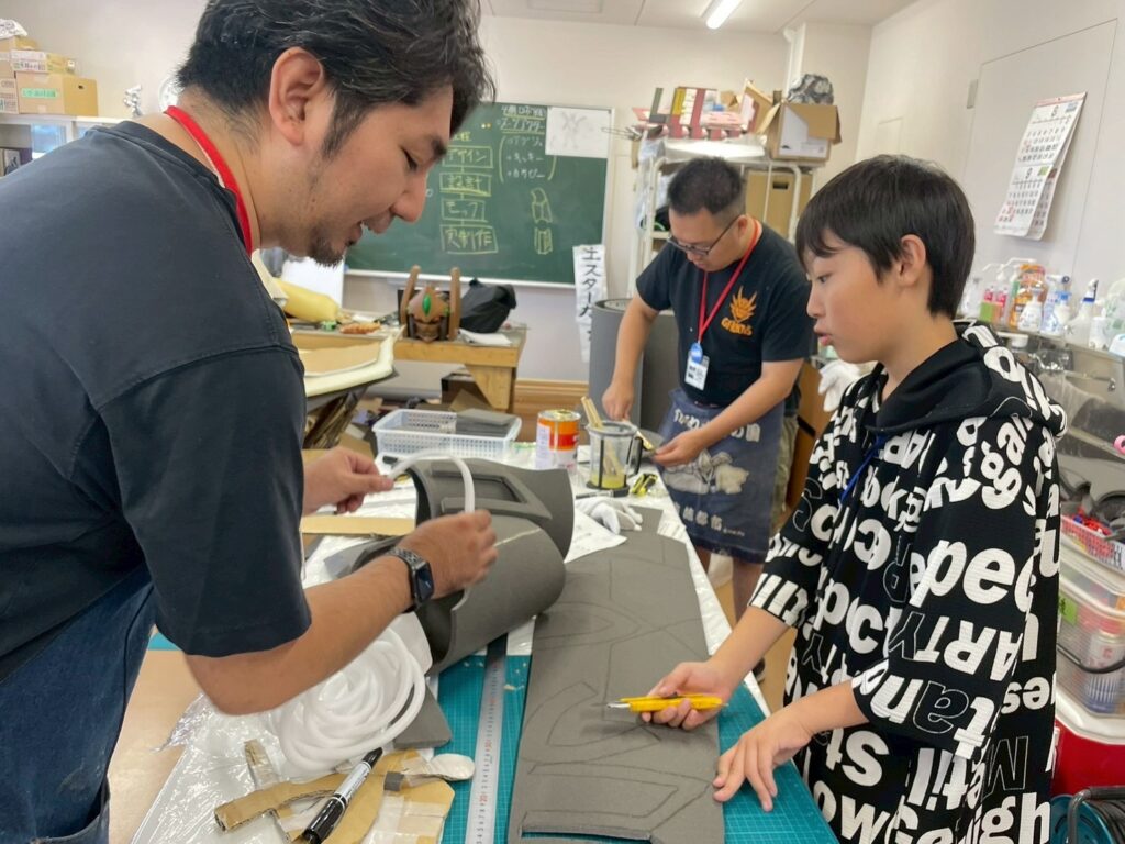 作業場の机で男性と男の子が発泡素材の大きな部材をカッターで慎重に加工し、その奥で男性が塗装や組立作業を進めている様子を写した写真