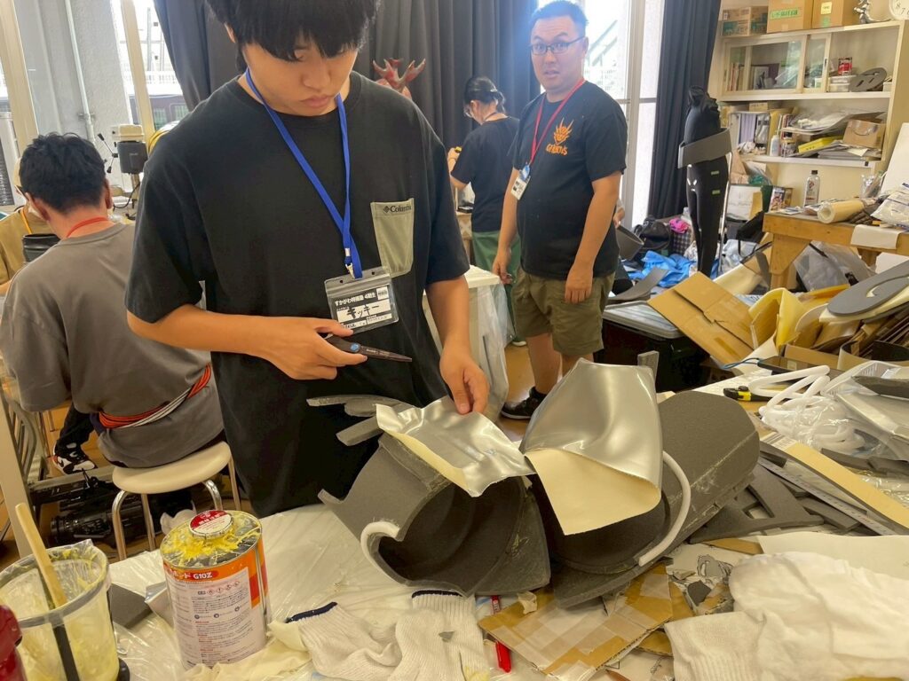 作業場の机で参加者が発泡素材の大きな部材に銀色シートを丁寧に貼り付けながら工作を進める写真