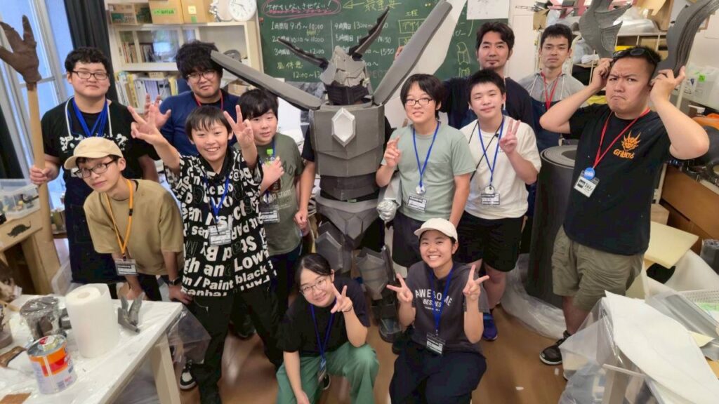 工作室の雑多な机や黒板を背景に大型ロボ風の立体作品を中心に多人数が工具や素材に囲まれ楽しげに集合する写真