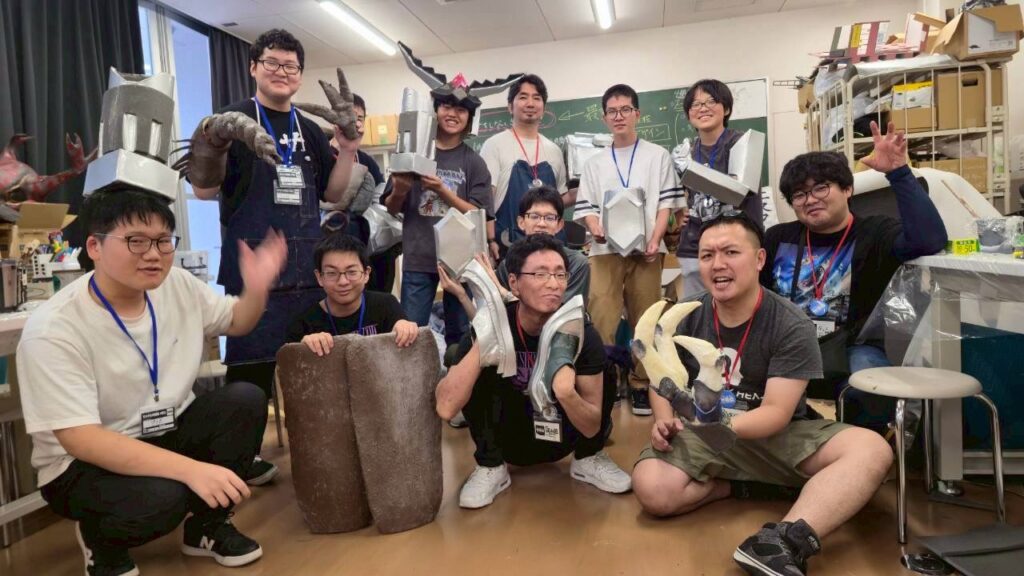 工作室で多数の塾生が自作の大型アーマー風造形物や道具を囲んでにぎやかに記念撮影している様子の写真