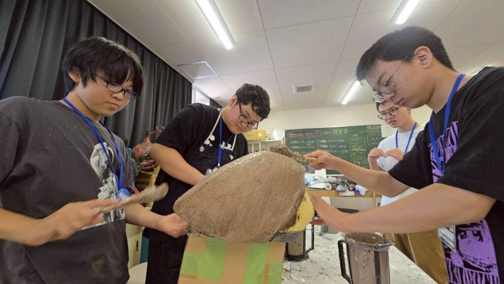 作業スペースで工具や材料が並ぶ中、複数の人が大きな球状の造形物に粘土を丁寧に塗り重ね形状を整えている写真