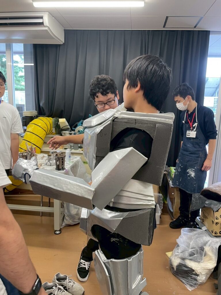 作業場で銀色と灰色のロボット風装甲を身に着けた塾生が腕部の可動を確認している様子を写した写真