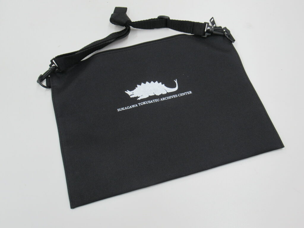 白地に置かれた黒いサコッシュが中央にあり、背中に鋭い突起を持ち、頭部から角のようなものが生えた怪獣のロゴと英字が刺繍されている写真