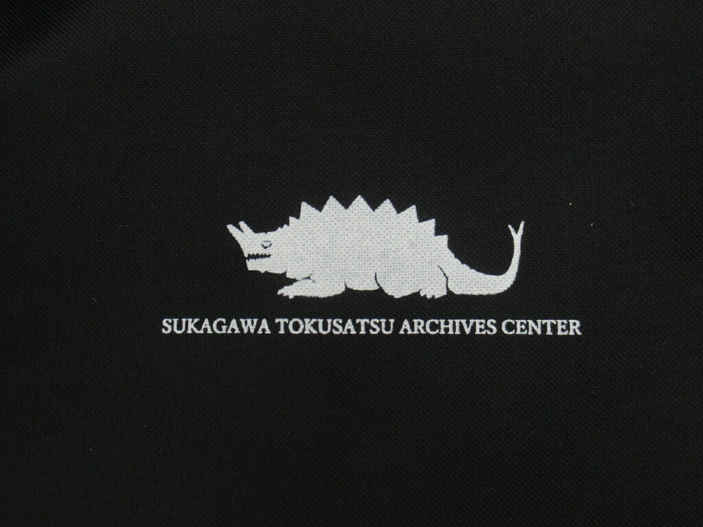 黒地に白い背中に鋭い突起を持ち、頭部から角のようなものが生えた怪獣のシルエットと“SUKAGAWA TOKUSATSU ARCHIVES CENTER”の文字が中央に配置されたデザインを大きく写した写真