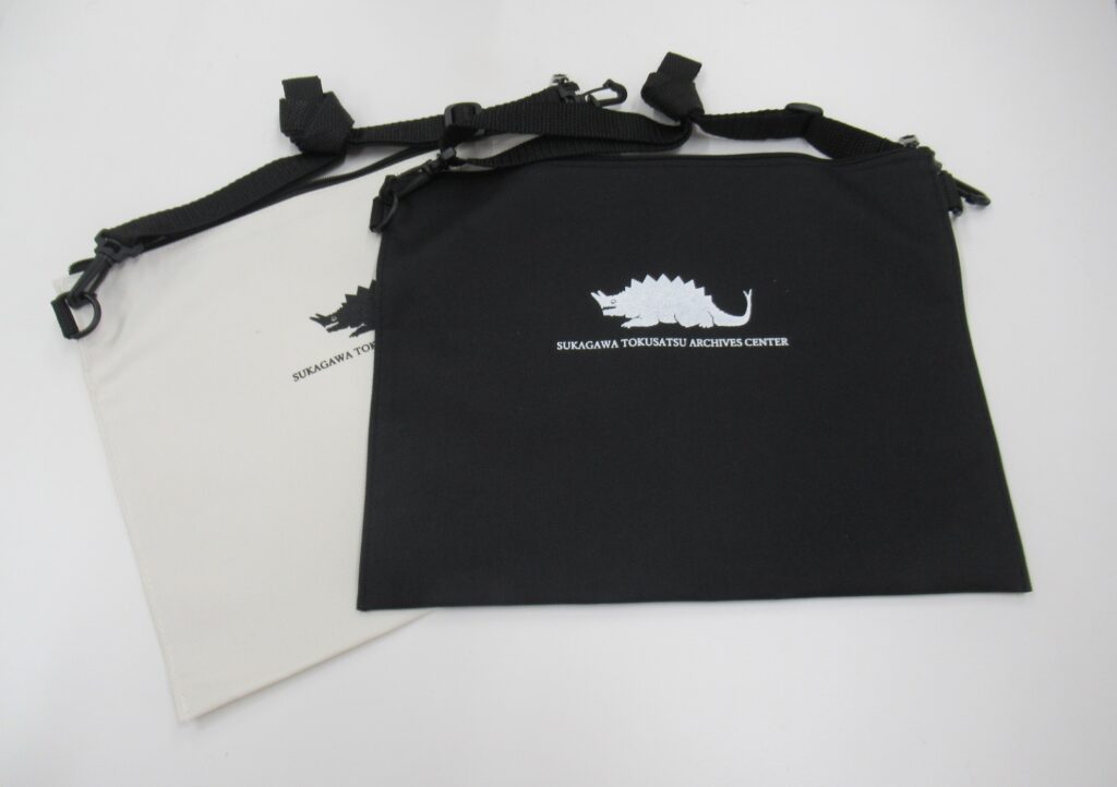 白い背景上で黒と生成りのサコッシュが重ねて置かれ、中央に背中に鋭い突起を持ち、頭部から角のようなものが生えた怪獣のロゴと文字が配された様子を写した写真