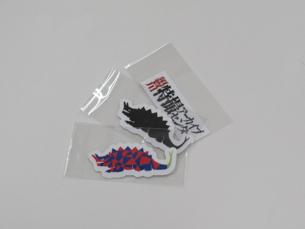 透明の袋に入った特撮アーカイブセンターの文字シールと黒色および多色の背中に鋭い突起を持ち、頭部から角のようなものが生えた怪獣のシールが重ねて置かれた様子を写した写真