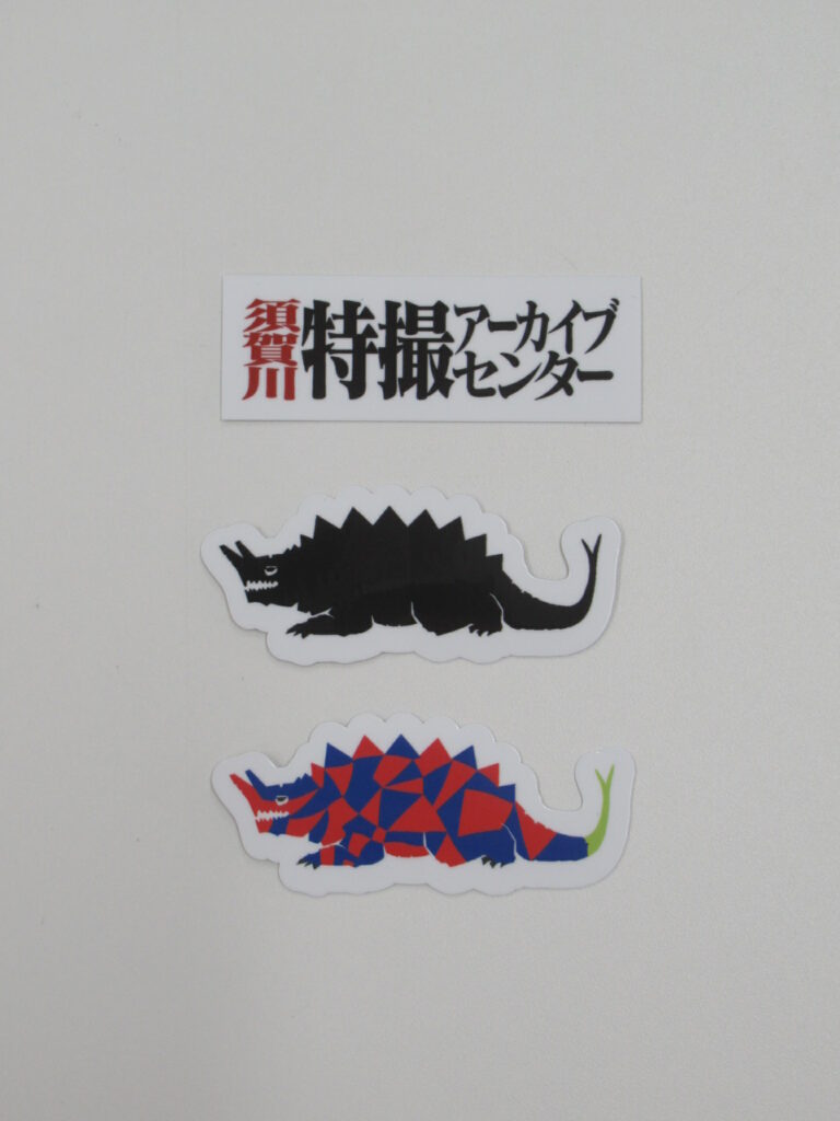 白い背景上に特撮アーカイブセンターの文字シールと黒色および多色の中央に背中に鋭い突起を持ち、頭部から角のようなものが生えた怪獣のシールが整然と並ぶ写真
