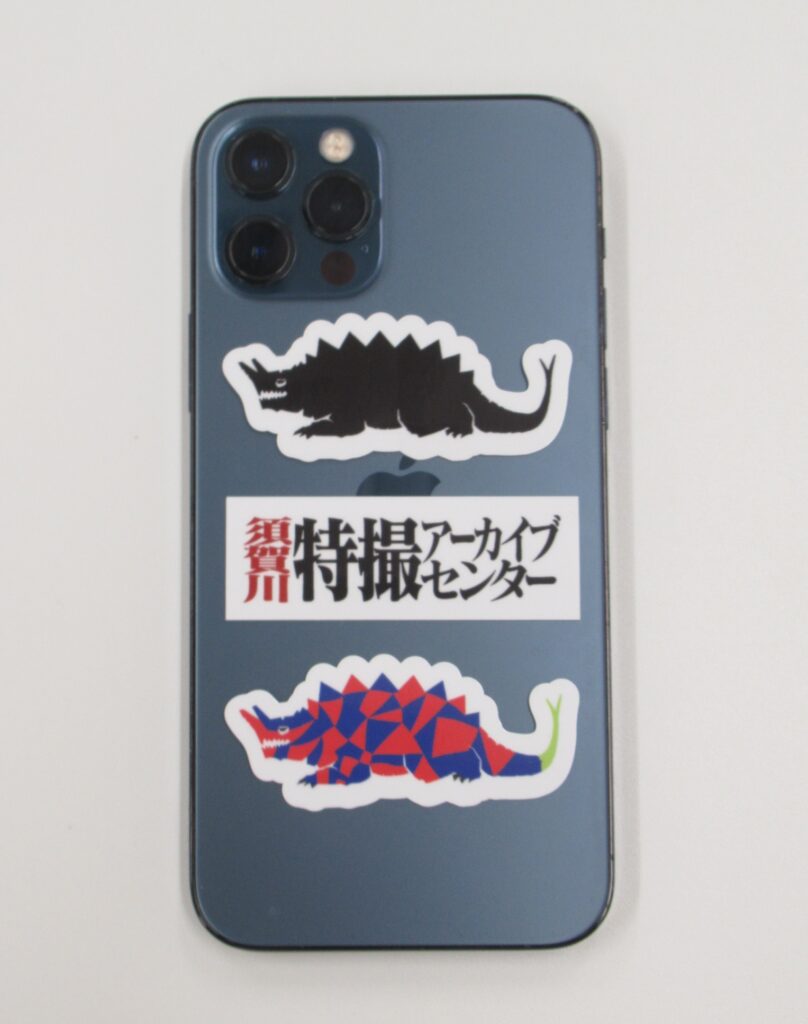 青いスマートフォン背面に黒と多色の背中に鋭い突起を持ち、頭部から角のようなものが生えた怪獣のシールと特撮アーカイブセンターの文字シールが縦に並んで貼られている様子を写した写真