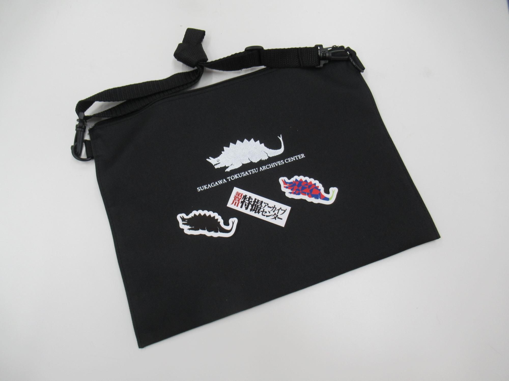 平らな白い背景上に置かれた黒いサコッシュに白い背中に鋭い突起を持ち、頭部から角のようなものが生えた怪獣のロゴとカラフルなシールが配置された様子を写した写真