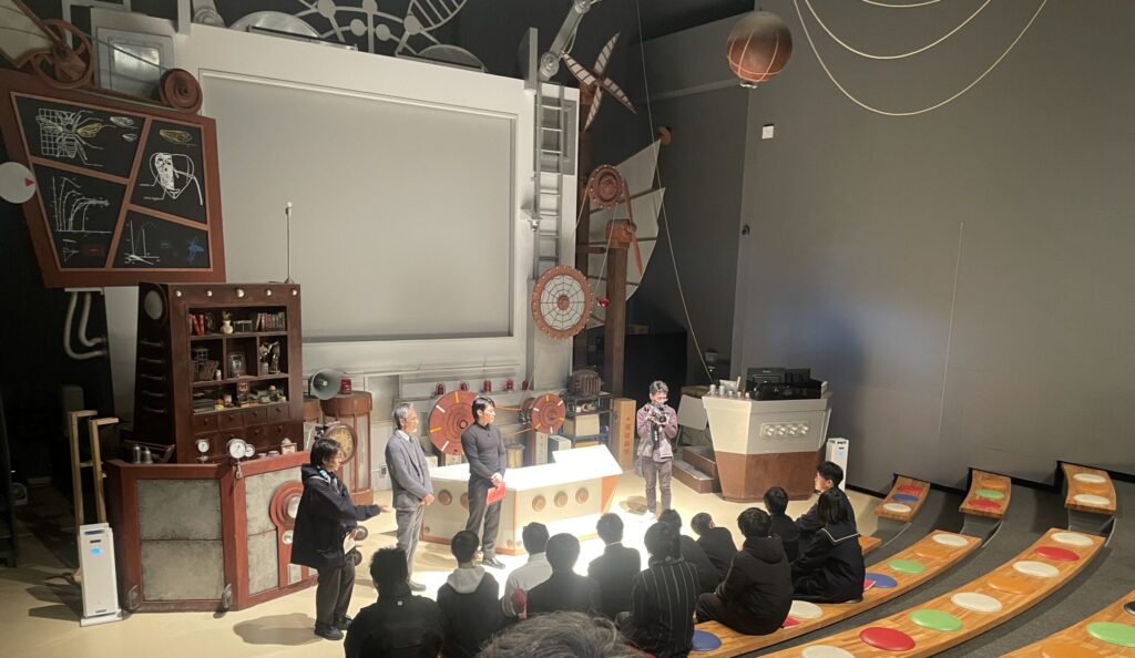 装飾された大型セット前で数名が立って説明を行い塾生が段状の席に座って真剣に話を聞く様子の写真