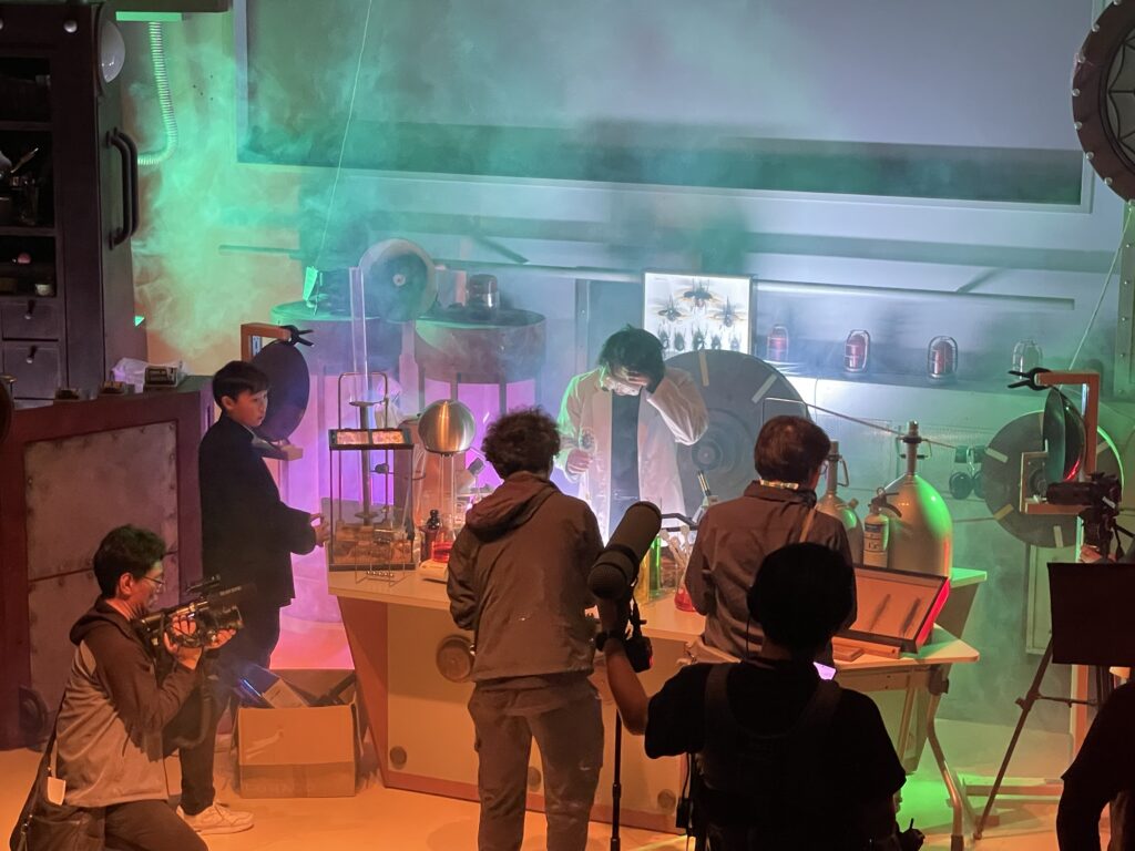 撮影セットの研究机の前で白衣姿の人物が装置を操作し周囲でスタッフがカメラや録音機材を構えて発光や煙の演出を伴う実験場面を撮影している様子の写真