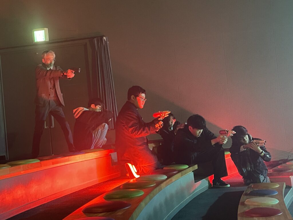 段状の席で複数の人物が赤い照明を浴びながら小道具の銃を構え、緊迫した場面を演じている様子の写真