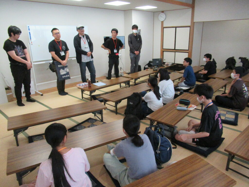 畳の部屋で、ホワイトボードの前に立つ5人の大人の話を、座卓に座った10人ほどの塾生が真剣な表情で聞いている写真