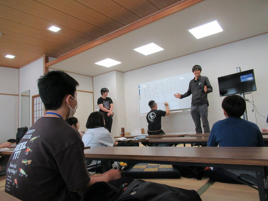 和室の研修会場で、一人の男性がホワイトボードの前で身振り手振りで説明を行い、もう一人の男性がその横で内容を書き留める様子を、長机に座った塾生が真剣に聞いており、その様子を後方から捉えた写真