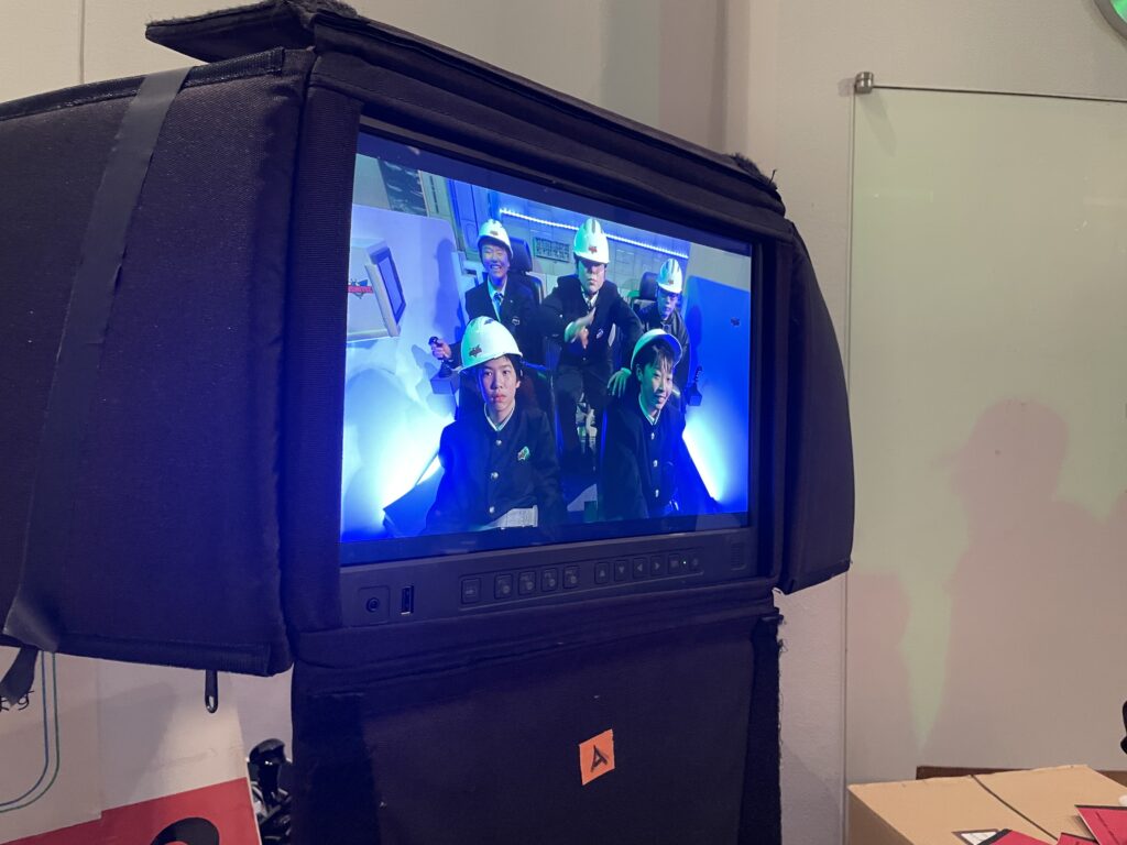 複数の塾生がヘルメットを着用して隊列を組み操縦席に座った様子が映し出されたモニターの写真