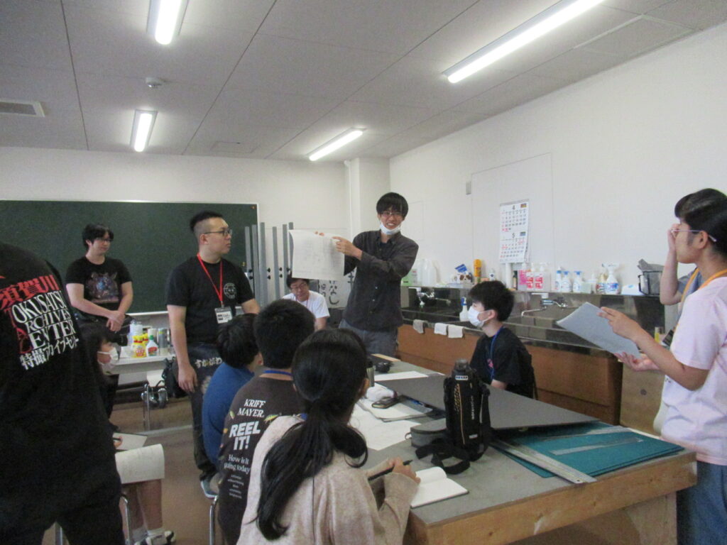 明るい作業室内で、講師の男性が図面の描かれた紙を掲げながら説明し、机を囲んで座る塾生がその手元を注視していたり、メモを取ったりしている写真