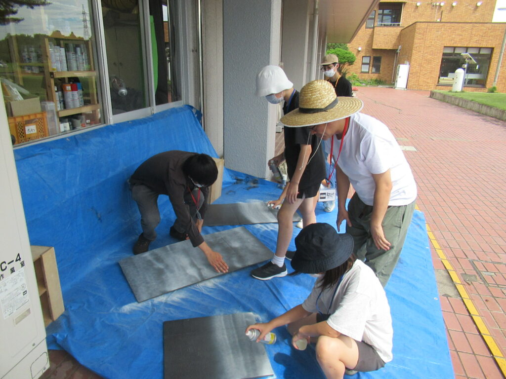 屋外の軒下に敷かれたブルーシートの上で、帽子を被った数人の塾生がスプレー缶を手に持ち、平らな板に灰色の塗料を吹き付けて着色作業を行い、それを大人のスタッフが傍らで見守りながら指導している制作風景の写真