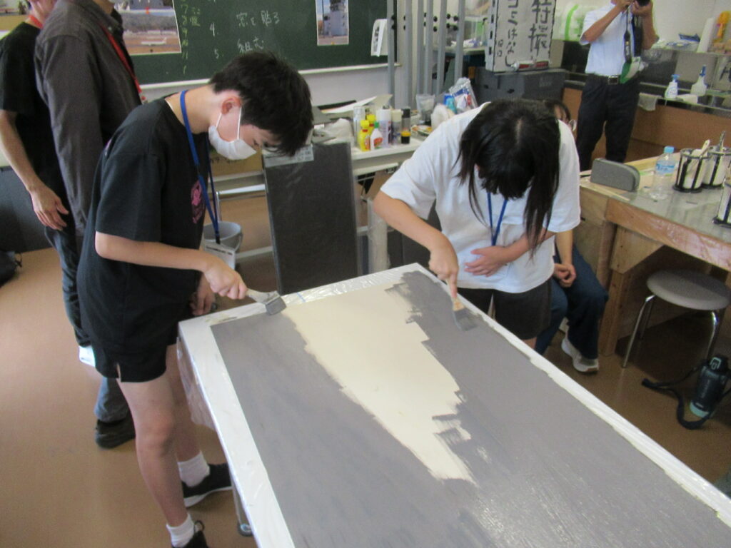 作業室内で、マスクを着用した2人の塾生が刷毛を手に持ち、机の上に広げた大きな白い板の両端から灰色の塗料を丁寧に塗り進めている作業風景の写真