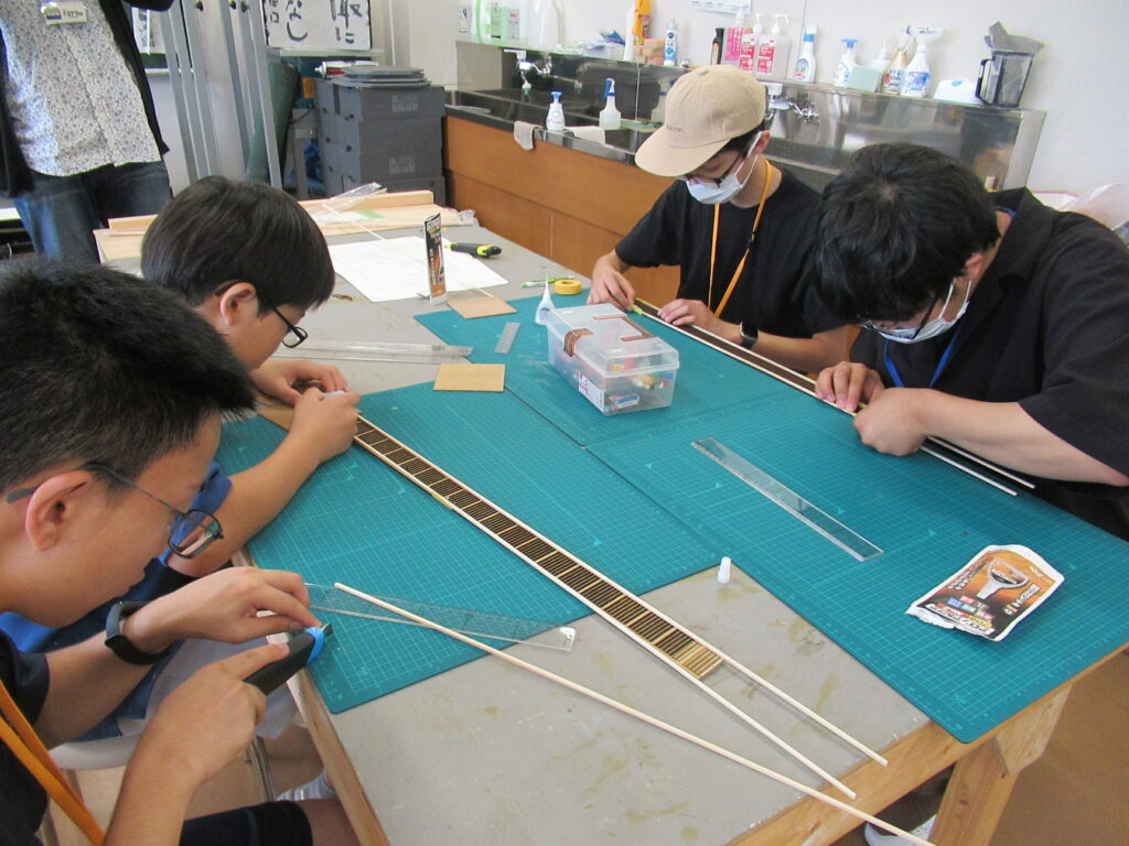 明るい作業室内で、カッターマットが敷かれた机を囲んだ4人の塾生が、定規やカッターを使いながら細長い木材やパーツを慎重に切り出し、ミニチュア模型の部品を組み立てる細かな作業に集中している制作風景の写真