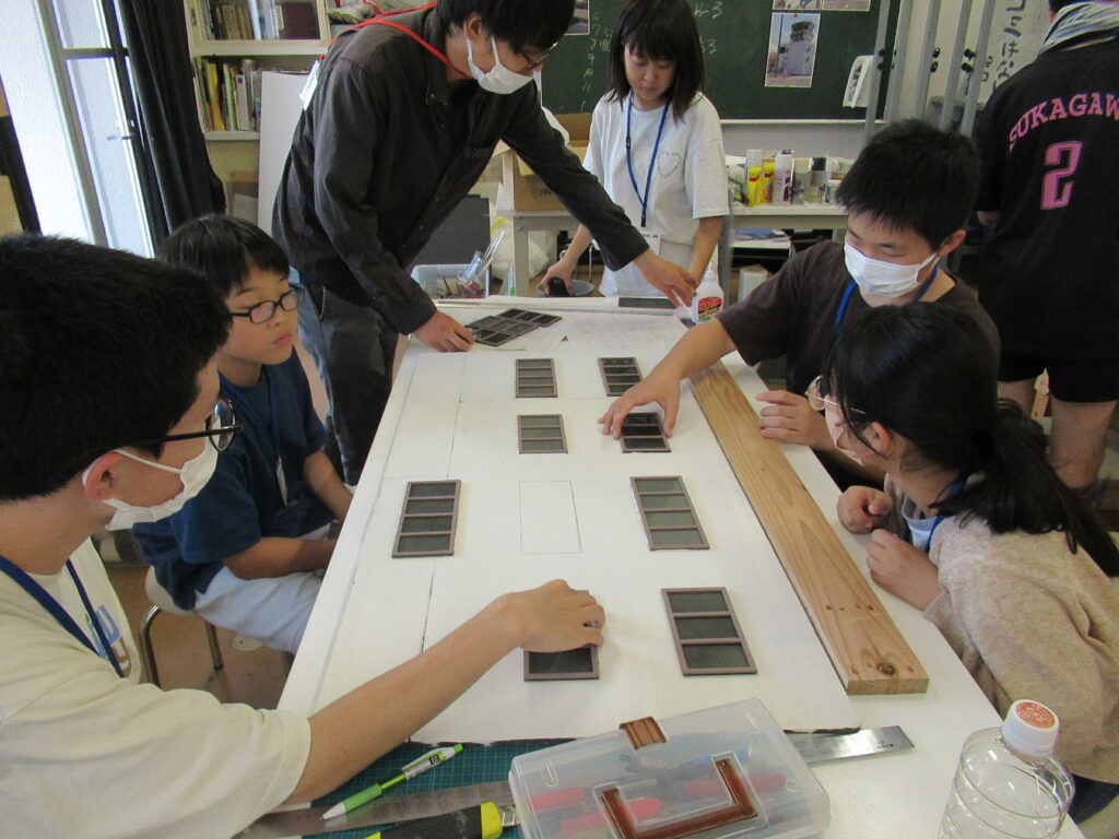 明るい作業室内で、複数の塾生が大きな白いボードの上に窓枠のようなパーツを等間隔に配置し、講師がその位置を指し示しながら微調整の指示を出している、建物のミニチュア模型を共同で制作する工程を捉えた写真