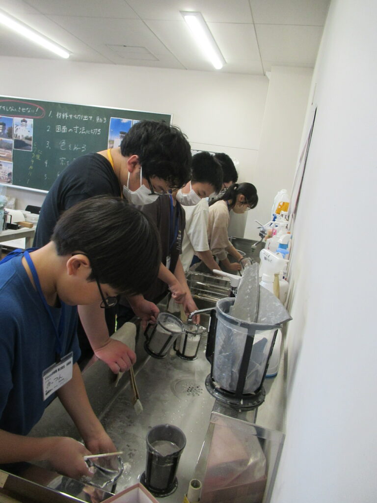 明るい作業室の流し台で、制作作業を終えた数人の塾生が、塗料の付いたカップや刷毛などの道具を水で丁寧に洗い流している写真