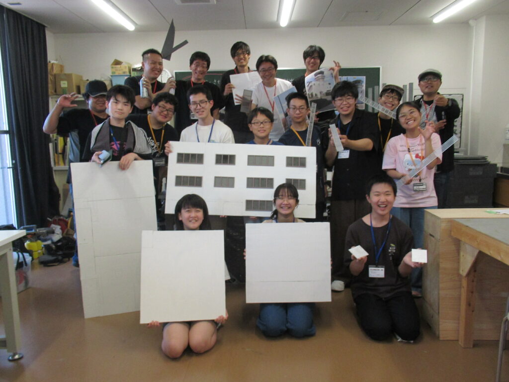 作業室内で、窓枠パーツが取り付けられた大きな白い外壁パネルを中央に掲げ、完成した各パーツを手にした塾生と講師陣が、晴れやかな笑顔の集合写真