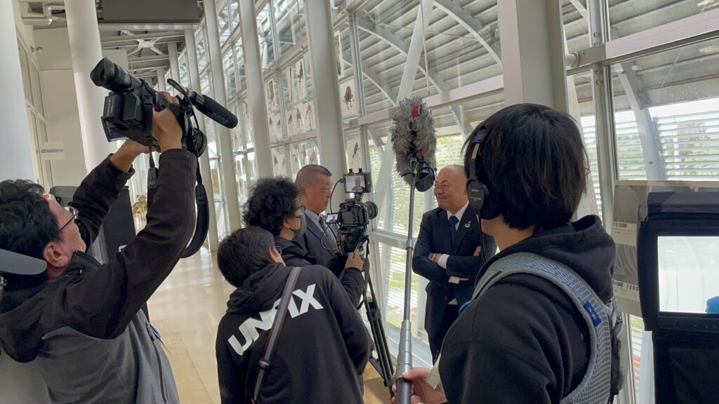 複数の撮影スタッフがカメラやブームマイクを構えて人物のインタビュー場面を至近距離で収録する様子の写真