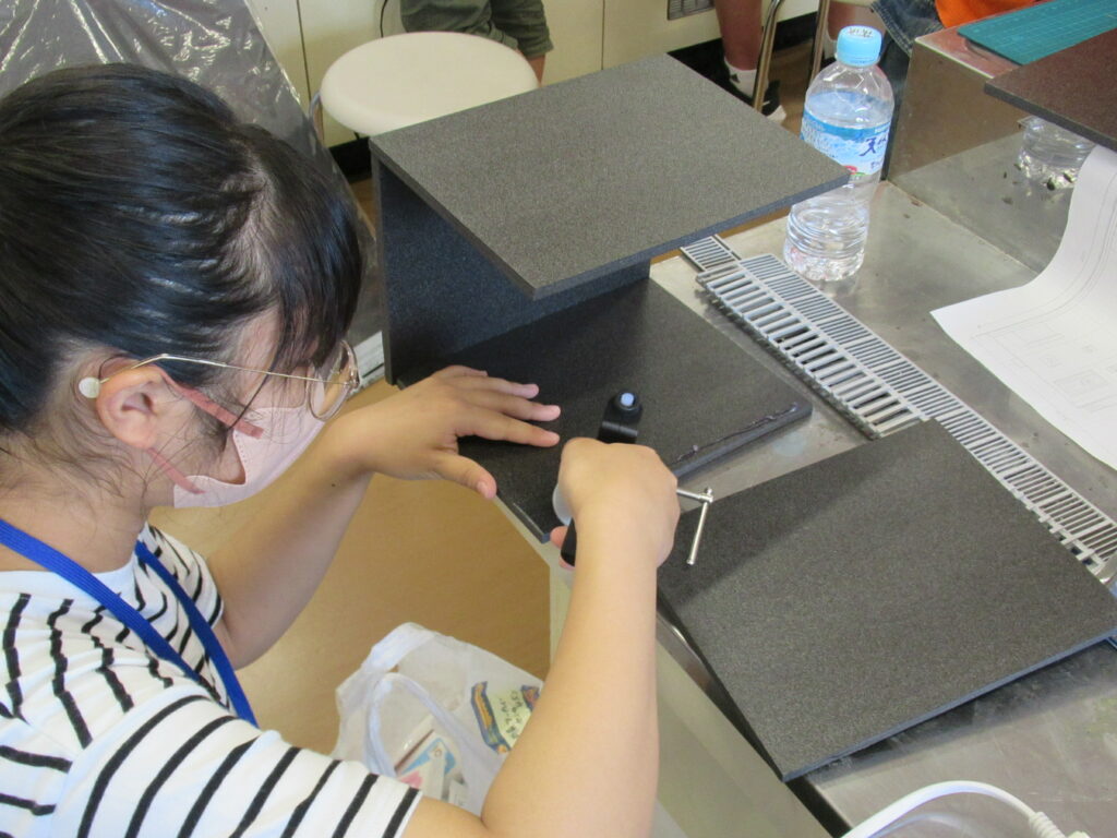 マスクと眼鏡を着用した塾生が、作業台の上で黒い板状のパーツを工具を使って慎重に組み立てている写真