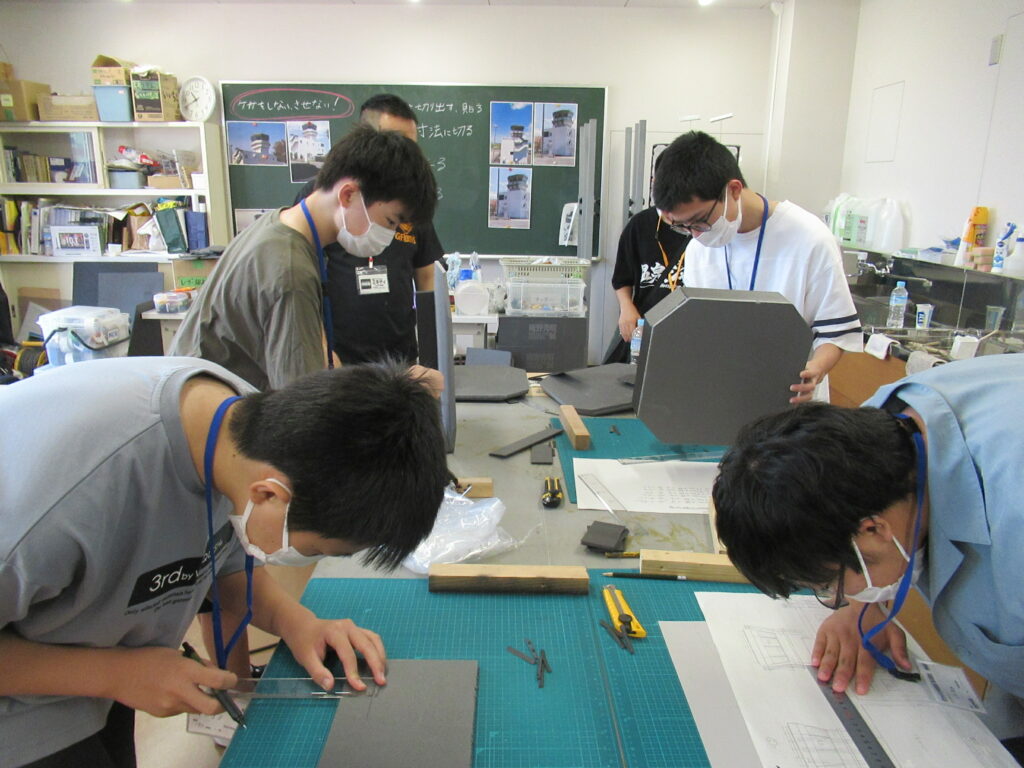 明るい作業室内で、マスクを着用した数人の塾生が、カッターマットが敷かれた机に向かい、図面を確認しながら定規やカッターを使って灰色のボードを正確な形に切り出している写真
