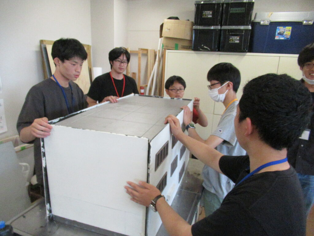 屋内の作業場で、数人の若い男性たちが真剣な表情を浮かべながら、窓の装飾が施された大きな白い建物模型の壁面を慎重に支えて組み立てたり、仕上がりを確認したりして制作に取り組んでいる様子を捉えた写真