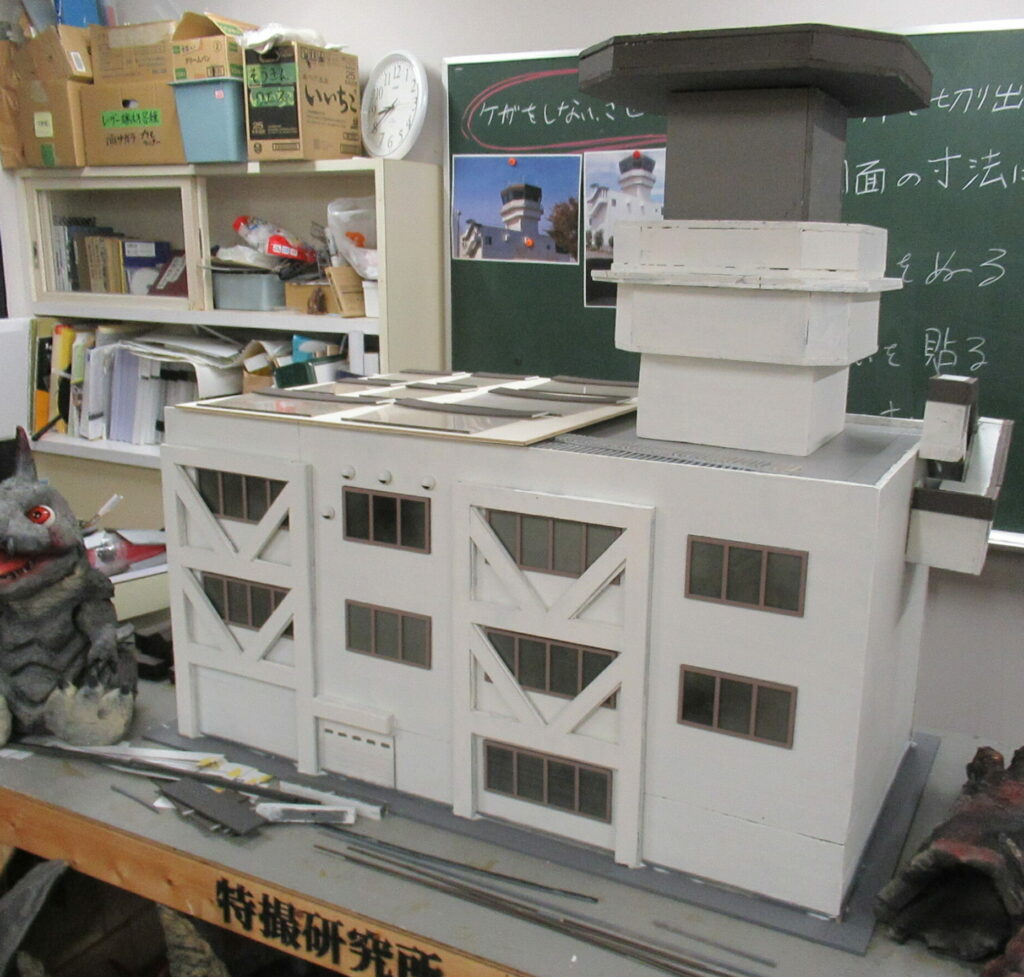 明るい作業室の机の上に、白壁に茶色の窓枠とV字型の補強構造が施された精巧な3階建てビルのミニチュア模型が完成し、屋上にはグレーの塔状パーツがそびえ立ち、傍らには特撮用の怪獣人形も置かれた模型の写真