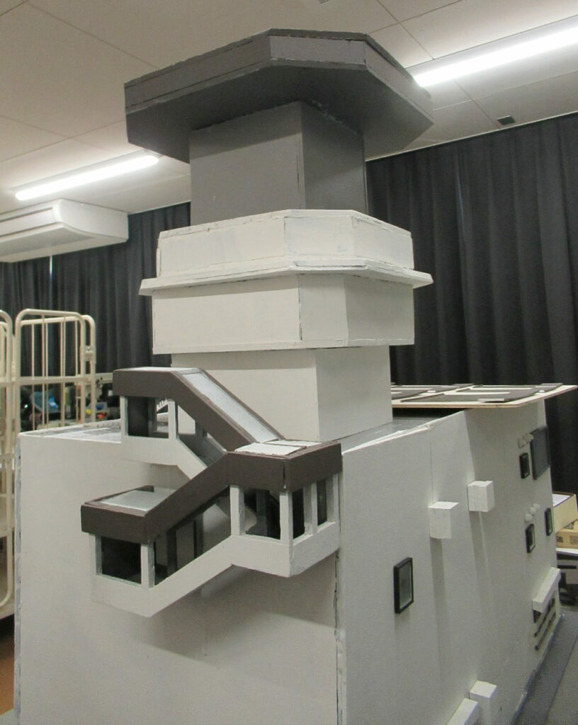 精巧に作られた3階建てビルのミニチュア模型が置かれ、建物の側面に階段が取り付けられ、屋上にはグレーの多角形の塔がそびえ立つ模型を近くから見た写真