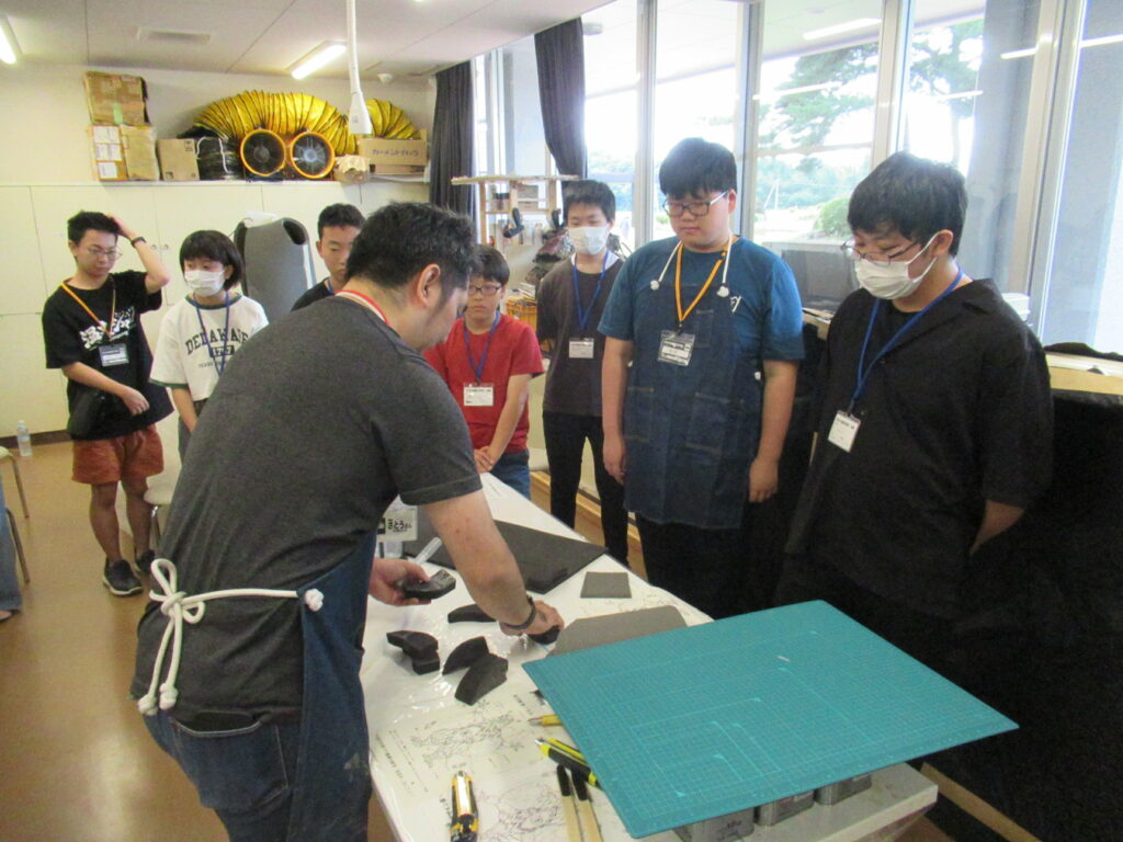 作業室内で、エプロン姿の講師が机の上に並べた黒いスポンジ状のパーツを使いながら成形しており、7人の塾生が真剣な眼差しで成形している様子を眺めている写真