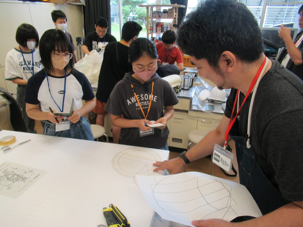 作業室内で、エプロンをつけた講師の男性が大きな白いボードに描かれた球体の図面を指し示しながら、メモを取る2人の塾生の写真