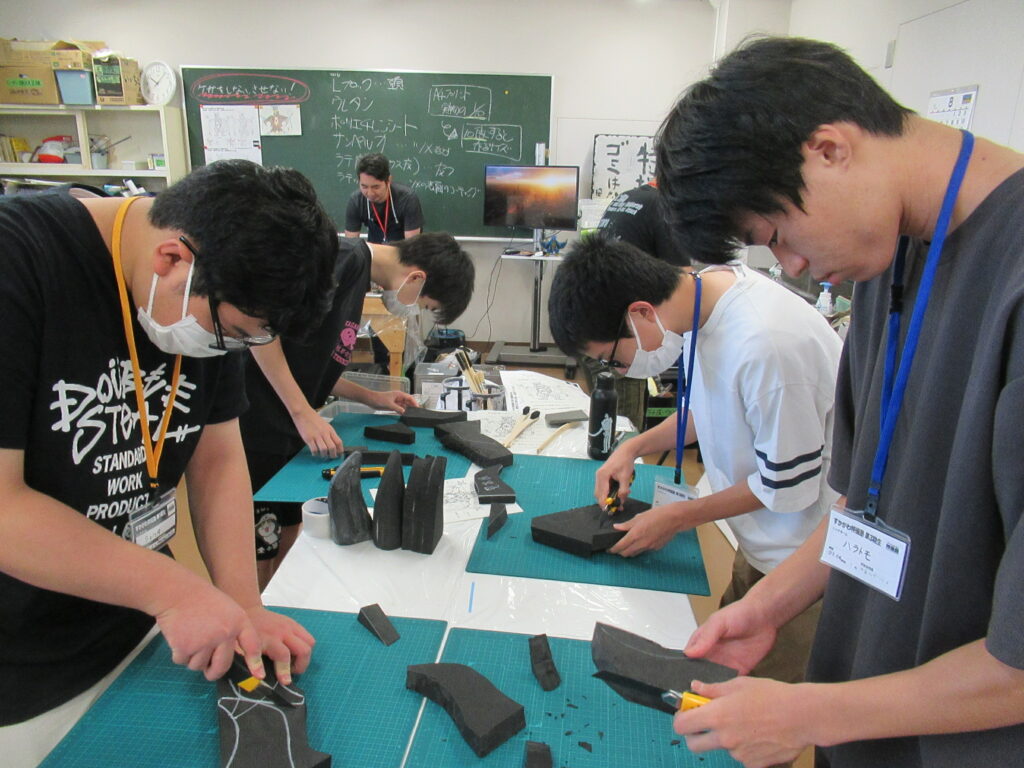 作業室内で4人の塾生が、カッターマットを敷いた机で黒いスポンジ状の造形材料を手に持ち、下書きの線に沿ってカッターで慎重に切り出している写真