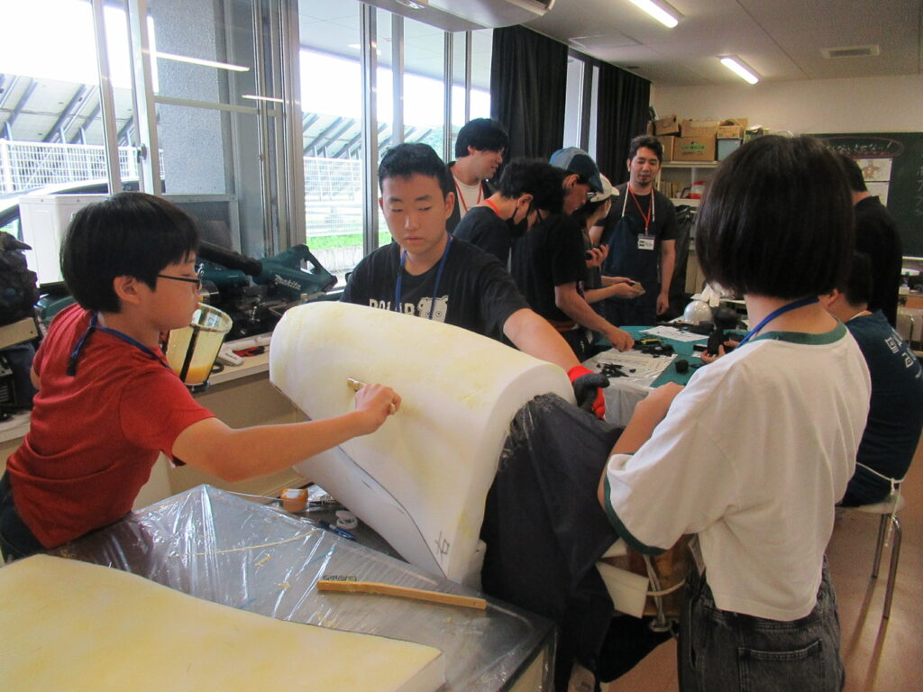 作業室内の大きな白いテーブルの上で、塾生が、巨大な円筒形の白いスポンジ状パーツを支えながらその表面に刷毛で黄色い液体を塗り広げている写真