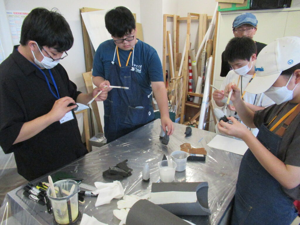作業室内の大きな白いテーブルを囲んだ4人の塾生が、黒いスポンジ状のパーツを手に持ち、刷毛を使って白い塗料や接着剤を表面に丁寧に塗り広げている写真