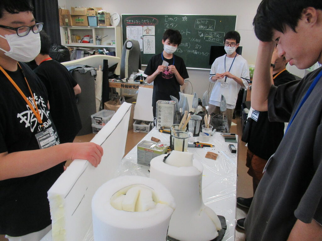 作業室内で、塾生がビニールを敷いた机の上に置かれた円筒形や半球状の白い大型造形パーツを囲み、全体の進捗を眺めている写真